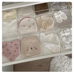 Może przedstawiać: Szuflada wypełniona ubrankami i akcesoriami dla niemowląt. Artykuły obejmują czapeczki, śliniaki, kapelusze i rękawiczki w różnych odcieniach bieli, kremu i różu. Niektóre przedmioty mają kwiatowe hafty lub koronkowe detale. Szuflada jest podzielona na przezroczyste przegródki.