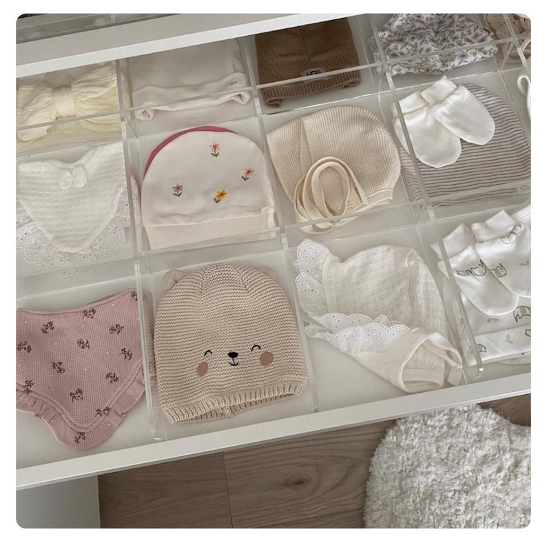 Puede incluir: Un organizador de cajones con varios art&iacute;culos para beb&eacute;s, incluyendo gorros, baberos y guantes. Los art&iacute;culos est&aacute;n organizados en compartimentos de pl&aacute;stico transparente. El caj&oacute;n est&aacute; hecho de madera blanca.