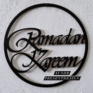 May include: Black metal circle with the words "Ramadan Kareem" written in a decorative script. The words are cut out of the metal and the circle is hanging on a white wall. The text "AUNDB PRESENTDESIGN" is also visible at the bottom of the circle.