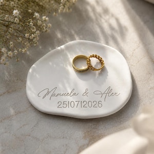Könnte beinhalten: Eine weiße, organisch geformte Schmuckschale aus Keramik mit der Gravur der Namen Manuela & Alex und dem Datum 25072026. Zwei goldfarbene Metallringe, ein schlichter Ring und ein Ring im Kettenglied-Design, liegen auf der glatten, matten Oberfläche.