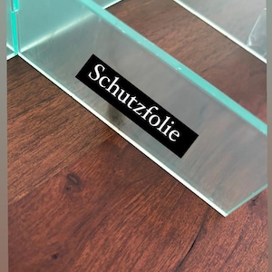 Puede incluir: Primer plano de un estante de vidrio transparente con una etiqueta negra que dice "Schutzfolie". El estante est&aacute; apoyado sobre una superficie de madera marr&oacute;n oscura.
