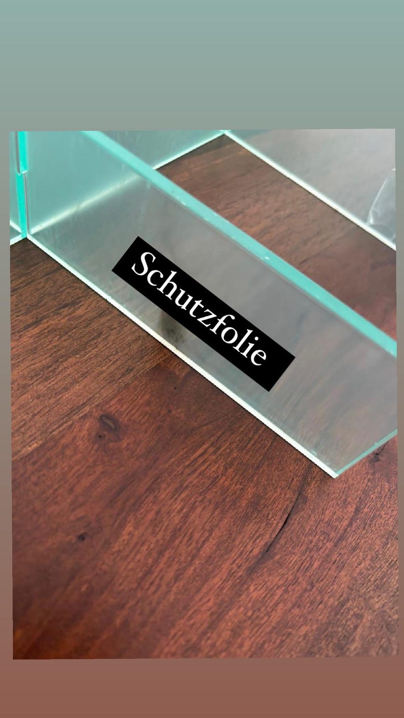 Puede incluir: Primer plano de un estante de vidrio transparente con una etiqueta negra que dice "Schutzfolie". El estante est&aacute; apoyado sobre una superficie de madera marr&oacute;n oscura.