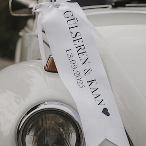 Puede incluir: Un coche blanco con una cinta decorativa blanca atada al capó. La cinta tiene los nombres "GÜLSEREN & KAAN" y la fecha "13.09.2025" impresos. Se ve el faro del coche.