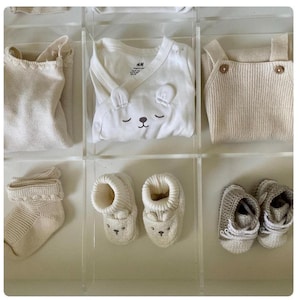 Peut inclure: Une collection de vêtements et d'accessoires pour bébés dans un organisateur en acrylique transparent. Les articles comprennent un onesie blanc avec un visage d'ours, des chaussons tricotés, des chaussettes et un pull. Les vêtements sont de tons neutres.