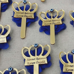 May include: Gold crown-shaped favors with blue fabric flowers and a blue evil eye charm. The favors have the text "Sünnet hatırası Karan Özçelik" written on them.