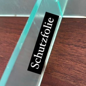Puede incluir: Primer plano de un estante de vidrio transparente con una etiqueta negra que dice "Schutzfolie". El estante est&aacute; sobre una superficie de madera.