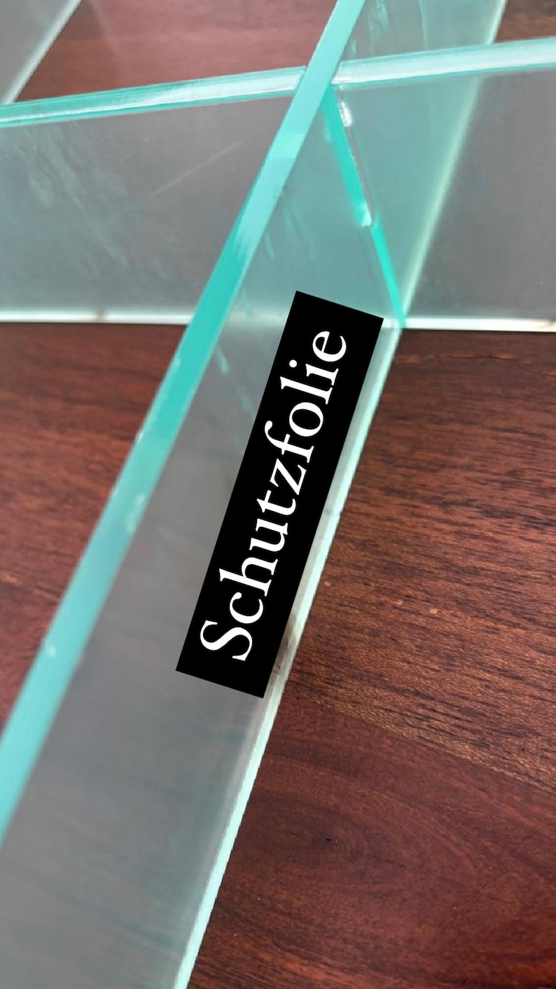 Puede incluir: Primer plano de un estante de vidrio transparente con una etiqueta negra que dice "Schutzfolie". El estante est&aacute; sobre una superficie de madera.