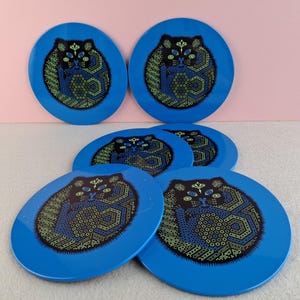 image shows a set of 6 Reginald Corfield black cat placemats, each is 16.5cm diameter circular metal & cork coasters with striking psychedelic design. The photos are taken against a stone and pink background.