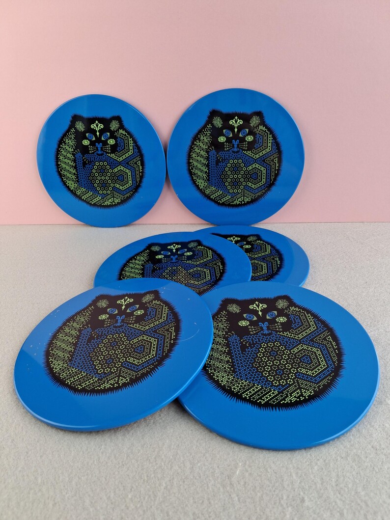 image shows a set of 6 Reginald Corfield black cat placemats, each is 16.5cm diameter circular metal & cork coasters with striking psychedelic design. The photos are taken against a stone and pink background.