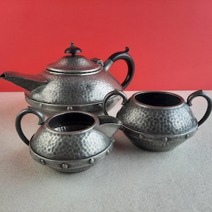 May include: A silver tea set with a hammered finish. The set includes a teapot, creamer, and sugar bowl. The teapot has a black handle.