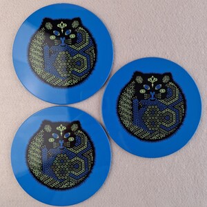 image shows a set of 6 Reginald Corfield black cat placemats, each is 16.5cm diameter circular metal & cork coasters with striking psychedelic design. The photos are taken against a stone and pink background.