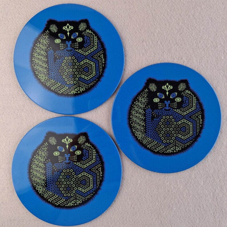 image shows a set of 6 Reginald Corfield black cat placemats, each is 16.5cm diameter circular metal & cork coasters with striking psychedelic design. The photos are taken against a stone and pink background.