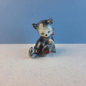 May include: A small, grey and white ceramic figurine of a cat with red berries on its collar. The cat is sitting with its paws tucked under its body.