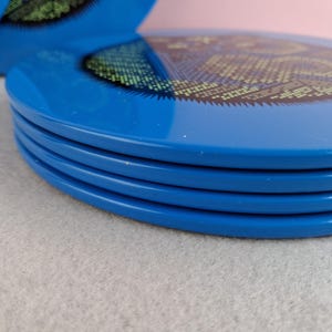 image shows a set of 6 Reginald Corfield black cat placemats, each is 16.5cm diameter circular metal & cork coasters with striking psychedelic design. The photos are taken against a stone and pink background.