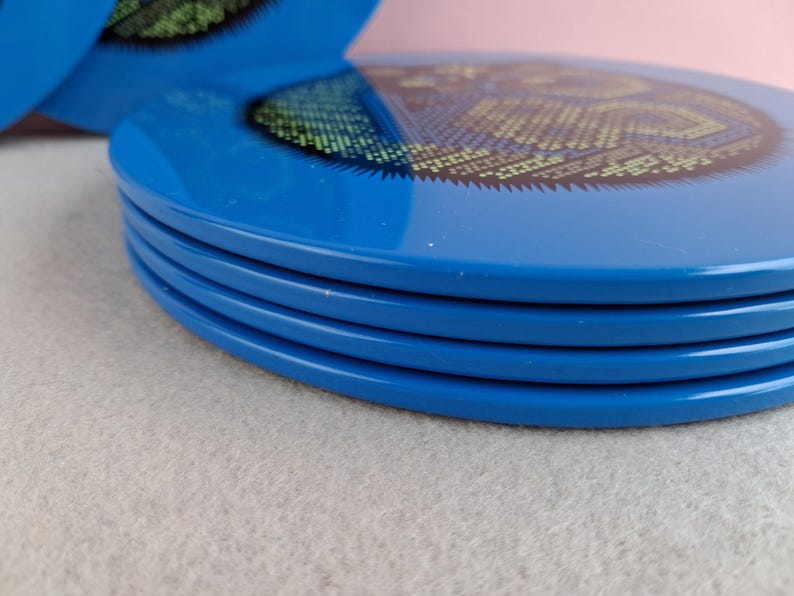 image shows a set of 6 Reginald Corfield black cat placemats, each is 16.5cm diameter circular metal & cork coasters with striking psychedelic design. The photos are taken against a stone and pink background.
