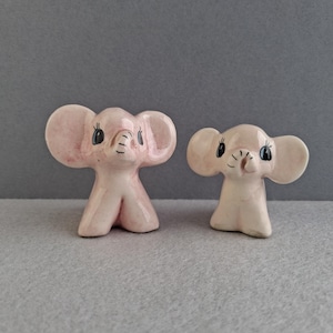 May include: Two small, pink ceramic elephant figurines with large ears and black eyes. The elephants have a vintage aesthetic and are displayed on a grey surface against a grey background. The figurines are likely decorative items.