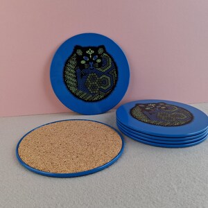 image shows a set of 6 Reginald Corfield black cat placemats, each is 16.5cm diameter circular metal & cork coasters with striking psychedelic design. The photos are taken against a stone and pink background.