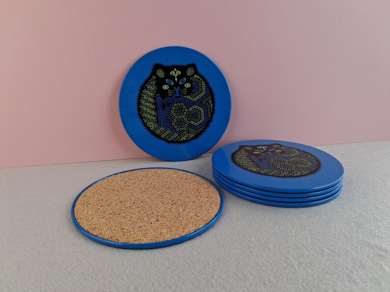 image shows a set of 6 Reginald Corfield black cat placemats, each is 16.5cm diameter circular metal & cork coasters with striking psychedelic design. The photos are taken against a stone and pink background.