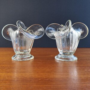 May include: Two clear glass vases with a distinctive design. Each vase features a flared top with a circular accent and a textured, patterned body. The vases are displayed on a wooden surface against a dark blue background.