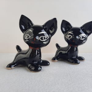 May include: Two black ceramic cat figurines with white eyes and black pupils. The cats are sitting with their heads turned to the side.