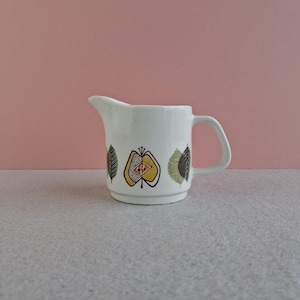 May include: White ceramic creamer with a handle and spout, decorated with colourful leaf and fruit designs. The creamer sits on a grey surface against a pink background. A vintage kitchenware item.