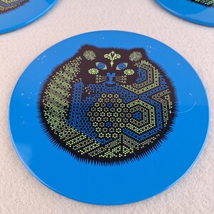 image shows a set of 6 Reginald Corfield black cat placemats, each is 16.5cm diameter circular metal & cork coasters with striking psychedelic design. The photos are taken against a stone and pink background.
