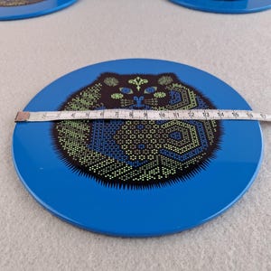 image shows a set of 6 Reginald Corfield black cat placemats, each is 16.5cm diameter circular metal & cork coasters with striking psychedelic design. The photos are taken against a stone and pink background.