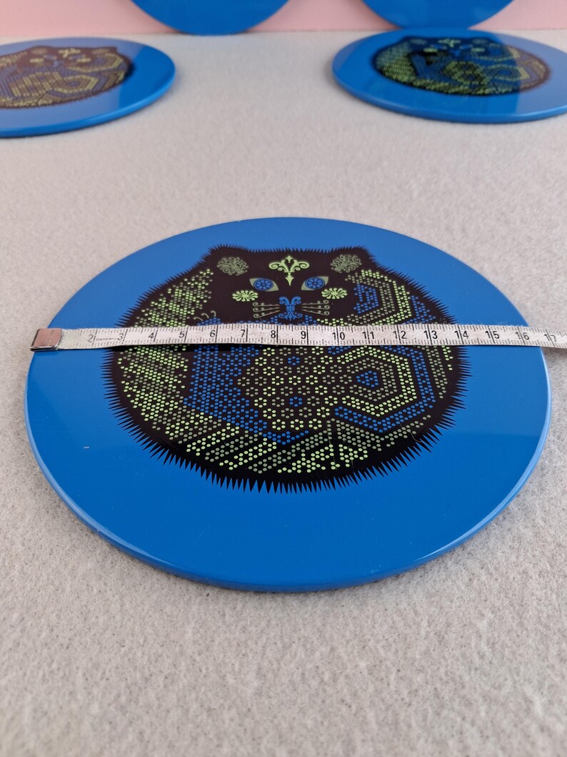 image shows a set of 6 Reginald Corfield black cat placemats, each is 16.5cm diameter circular metal & cork coasters with striking psychedelic design. The photos are taken against a stone and pink background.