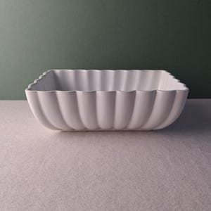 May include: A white, square ceramic bowl with a scalloped edge. The bowl is set against a muted green background and a light-coloured surface. The bowl's design features vertical, rounded ridges.