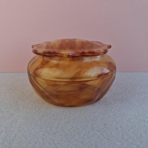 May include: A small, round, lidded jar with a marbled amber appearance. The jar has a rounded body and a lid with a scalloped edge. The jar is made of a translucent material, and the marbling creates a swirling pattern.