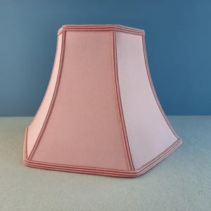 May include: A pink lampshade with a hexagonal shape and a scalloped edge. The lampshade has a subtle texture and a delicate, feminine design.