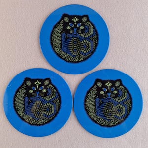 image shows a set of 6 Reginald Corfield black cat placemats, each is 16.5cm diameter circular metal & cork coasters with striking psychedelic design. The photos are taken against a stone and pink background.