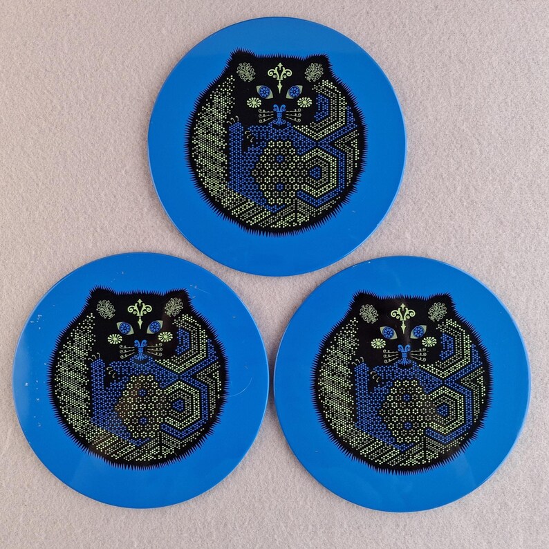 image shows a set of 6 Reginald Corfield black cat placemats, each is 16.5cm diameter circular metal & cork coasters with striking psychedelic design. The photos are taken against a stone and pink background.
