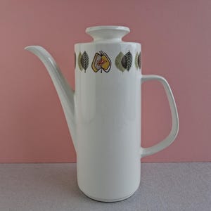 May include: A white ceramic coffee pot with a tall, cylindrical shape, a long spout, and a handle. The top has a decorative band of stylized leaves and fruit in green, yellow, and brown. The pot has a matching lid.