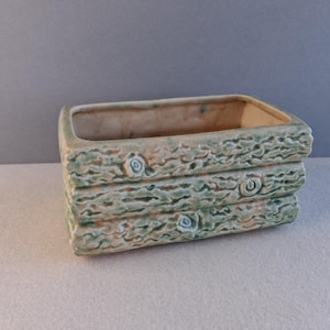 May include: Rectangular ceramic planter with a textured, stacked log design. The planter is light green with brown and white accents, and has a rectangular opening. Suitable for small plants or succulents.