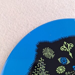image shows a set of 6 Reginald Corfield black cat placemats, each is 16.5cm diameter circular metal & cork coasters with striking psychedelic design. The photos are taken against a stone and pink background.