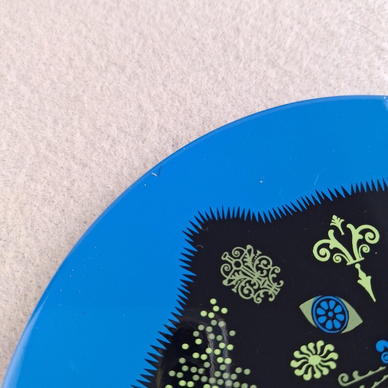 image shows a set of 6 Reginald Corfield black cat placemats, each is 16.5cm diameter circular metal & cork coasters with striking psychedelic design. The photos are taken against a stone and pink background.
