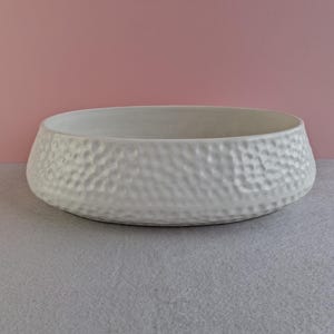 May include: A white bowl with a textured, hammered finish. The bowl has a low profile and sits on a grey surface, with a pink backdrop. This decorative bowl is suitable for serving or display.