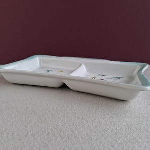 May include: A white, divided serving dish with a pale blue trim. The rectangular dish has a curved edge on each end. Small, colourful leaf designs are painted inside. The dish is on a light grey surface.