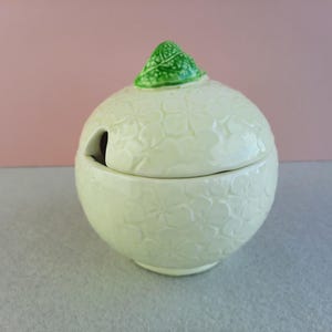 May include: A white ceramic sugar bowl with a green leaf lid. The bowl has a floral pattern and a small opening on the side.