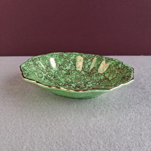May include: A small, oval dish with a scalloped edge. The dish is a vibrant green with a detailed floral pattern in gold and brown. The rim is trimmed with gold.