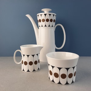 May include: A white ceramic coffee pot, creamer and sugar bowl with a black and brown geometric pattern. The pattern features circles and ovals.