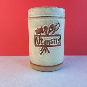 May include: A ceramic utensil holder with a light beige glaze and brown accents. The holder features a stylised design of utensils and the word "Utensils" in brown lettering.