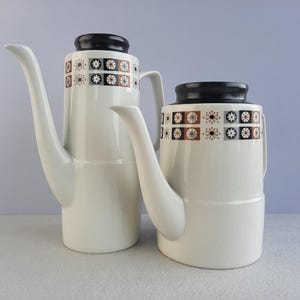 May include: Two white ceramic coffee pots with black and brown geometric patterns. The pots have a rounded body and a spout on the side.