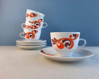 May include: A set of six white teacups with orange and brown floral designs. The cups are stacked on top of each other and are sitting on a white surface.