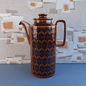 May include: A brown ceramic coffee pot with a lid and handle. The pot has a repeating pattern of dark blue and brown shapes. The spout is long and slender. The coffee pot is likely vintage, measuring approximately 25 cm in height.