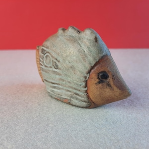 May include: A ceramic bird figurine with a light grey body and a brown beak. The bird has a textured surface and a small, dark eye. The background is a solid red.