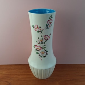 May include: A white ceramic vase with a flared shape, featuring a blue interior and a floral design of pink and white flowers with green leaves. The base has vertical ribbed detailing.