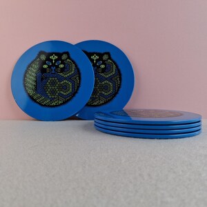 image shows a set of 6 Reginald Corfield black cat placemats, each is 16.5cm diameter circular metal & cork coasters with striking psychedelic design. The photos are taken against a stone and pink background.