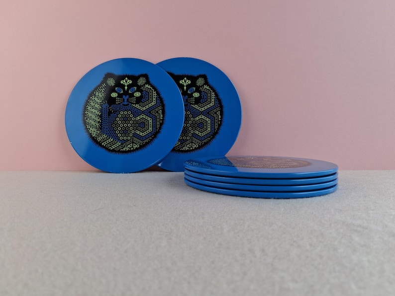 image shows a set of 6 Reginald Corfield black cat placemats, each is 16.5cm diameter circular metal & cork coasters with striking psychedelic design. The photos are taken against a stone and pink background.